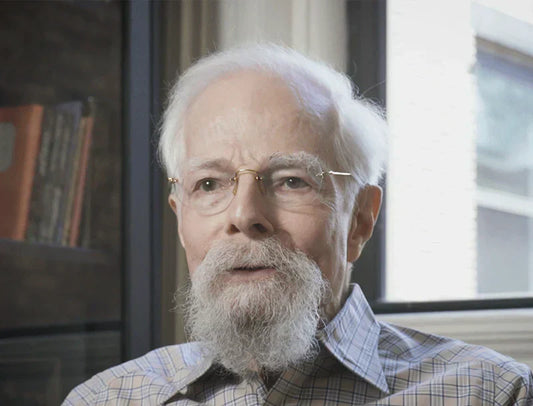 Elderly man with glasses and white beard indoors, bookshelf and window in background