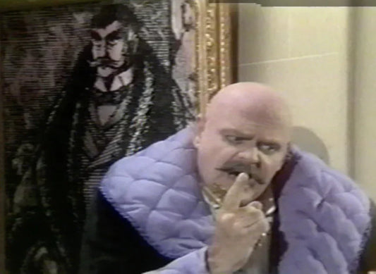 Bald man in a purple robe posing by framed Edward Gorey-style artwork, vintage setting