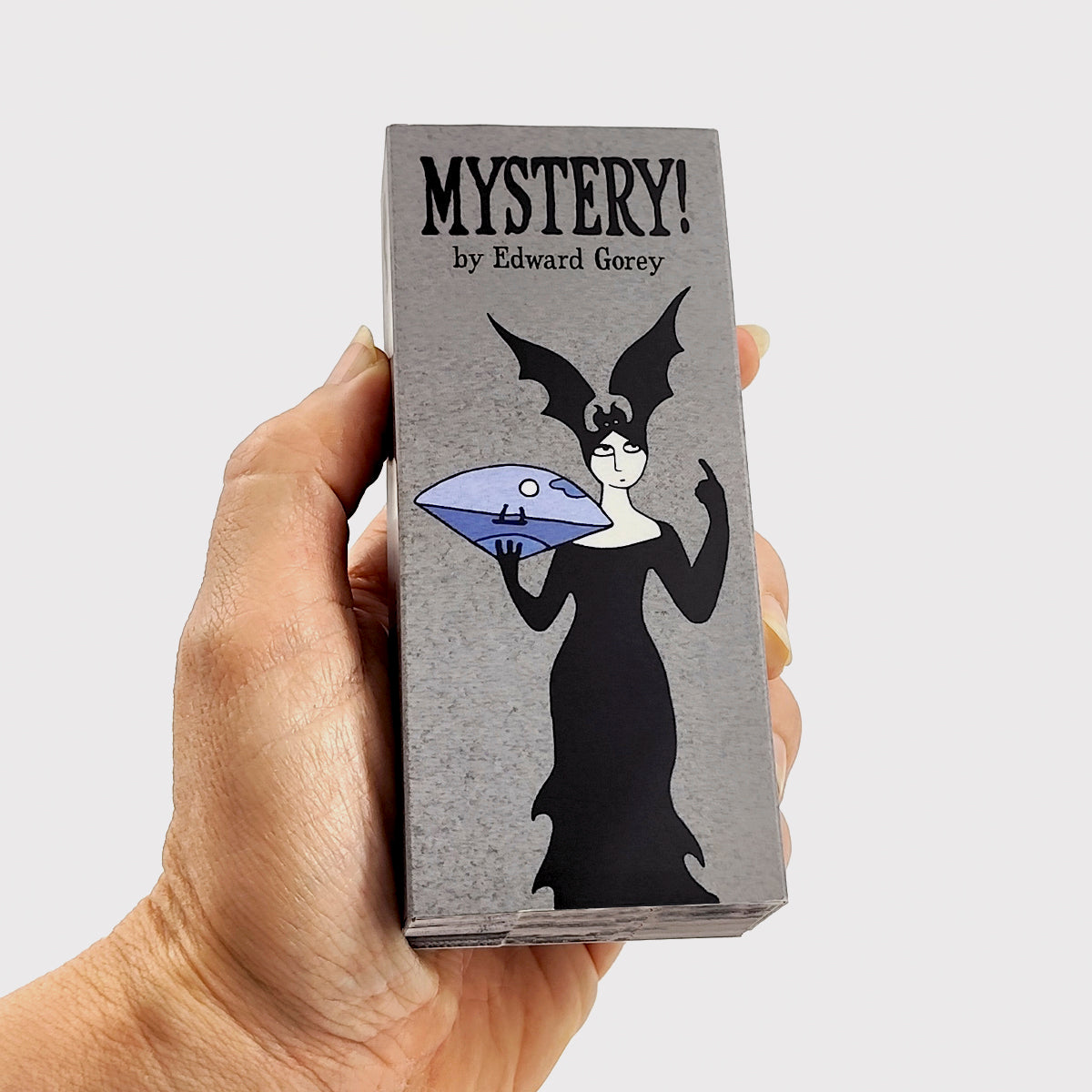 Hand holding Edward Gorey Mystery Flipbook with gothic woman illustration and bold title