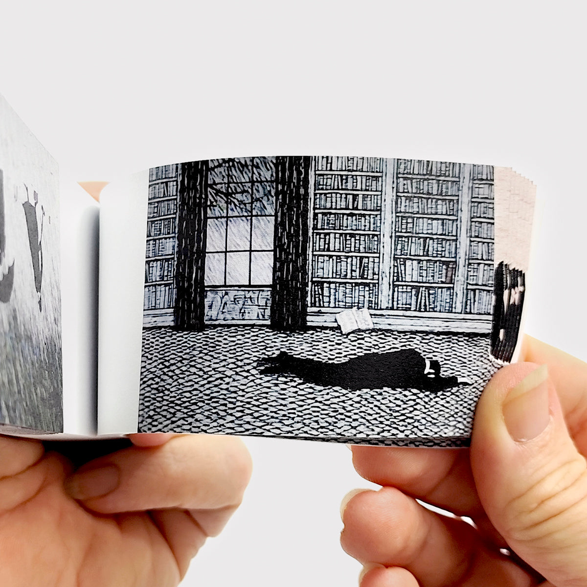 Edward Gorey Mystery Flipbook held open, showing a black and white illustration of a figure lying on a library floor with shelves of books in the background.