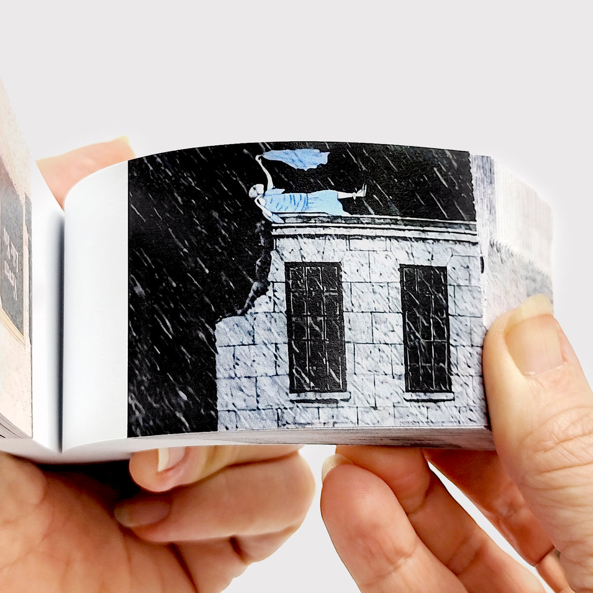 Hands flipping through Edward Gorey flipbook with illustrated figure on rooftop in the rain