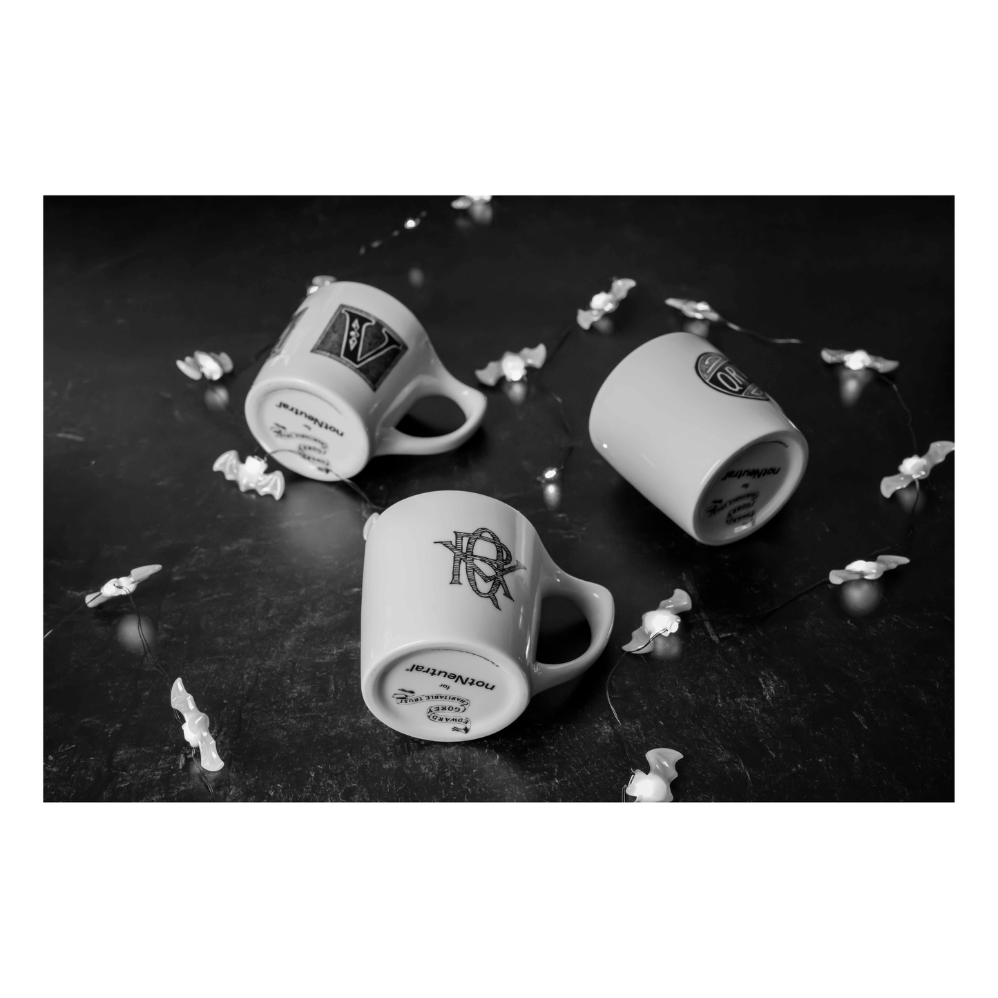 Edward Gorey Catastrophe mugs on dark surface with string lights, three upside down, gothic style.