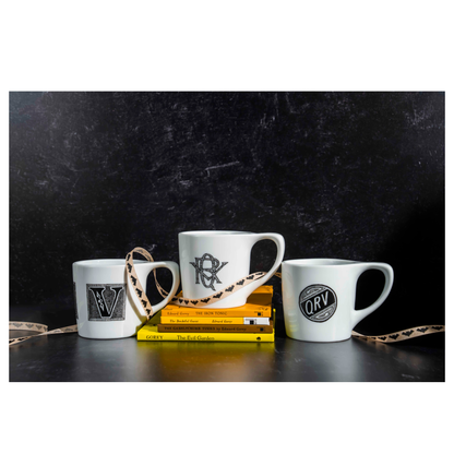 Three Edward Gorey mugs with monogram designs, stacked on Gorey books, black background
